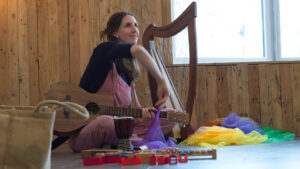 Laura sits with a small harp on the ground, wearing pink dungarees and smiling