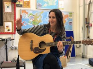 Laura sits in a chair, holding an acoustic guitar, wearing a stripey shirt